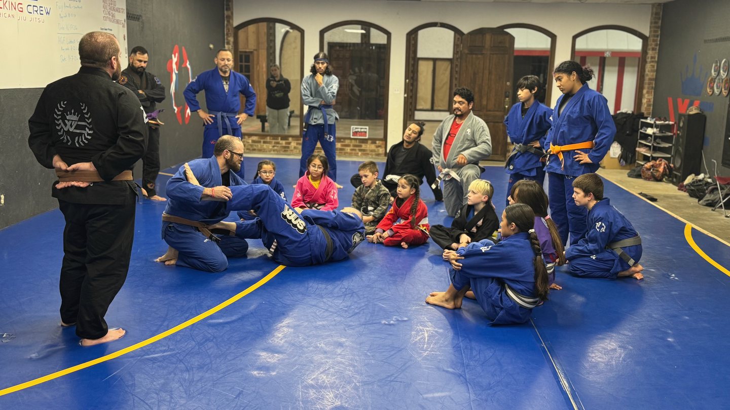 Adults and children watching a jiu jitsu demonstration on blue mats.