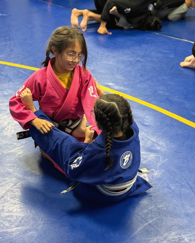 Two children in pink and blue gis practicing a partner drill on blue mats.